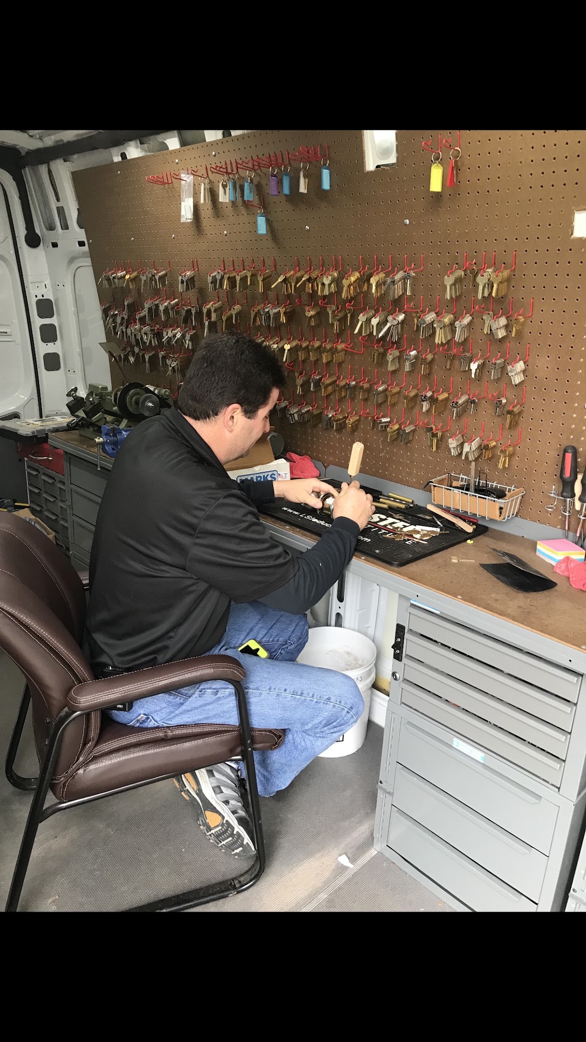A J & J locksmith cutting a key at the workbench inside the van, with a wall of key blanks behind